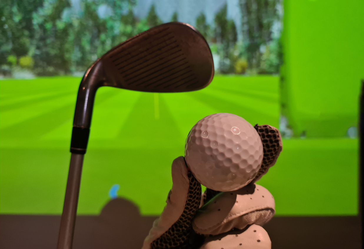 Golfer in glove holds golf ball and club indoors. Playing golf on a simulator
