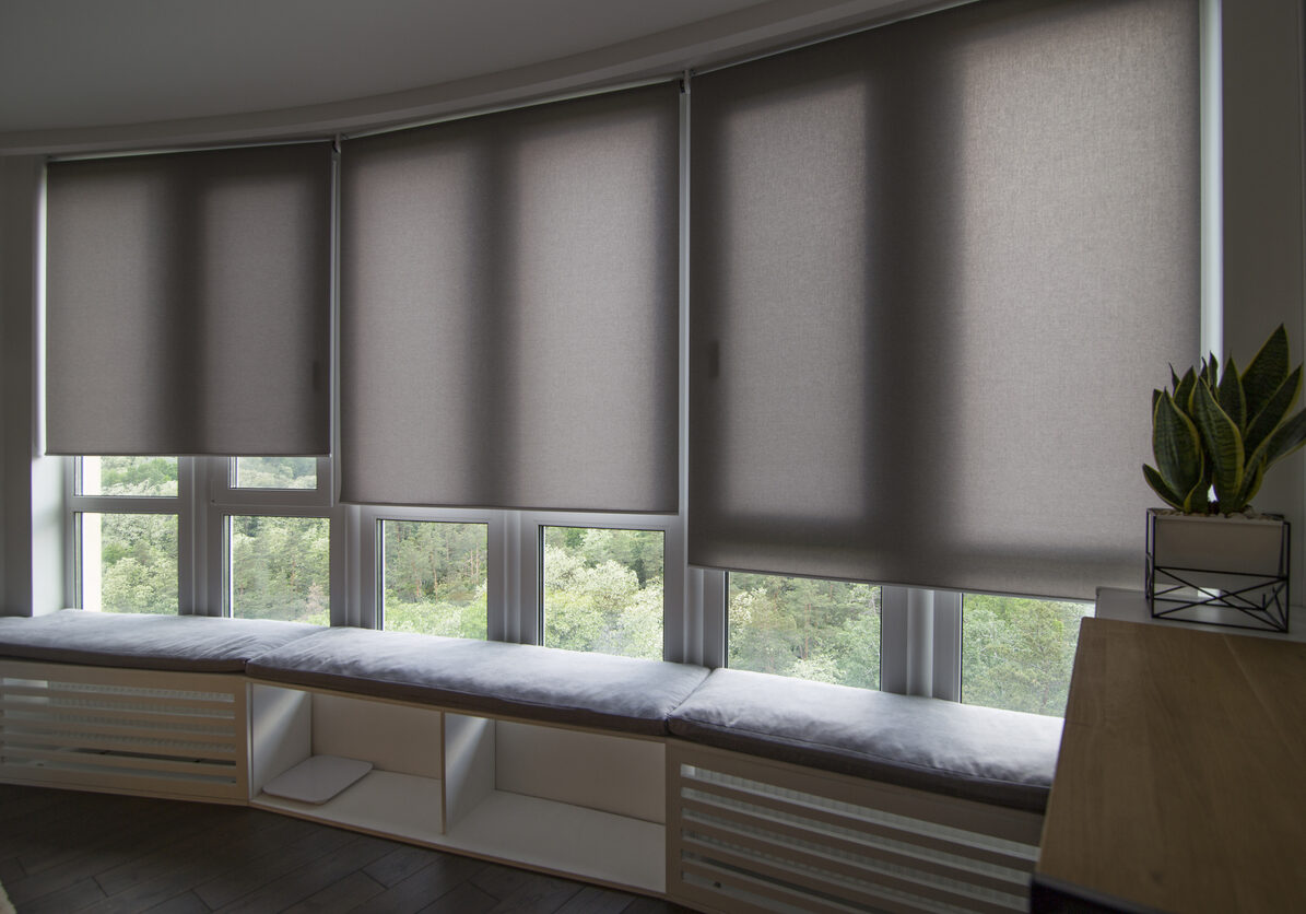 Motorized roller shades in the interior. Automatic roller blinds beige color on big glass windows. Home luxury curtaines are above the windosill with pillows. Electric blinds for home automation.