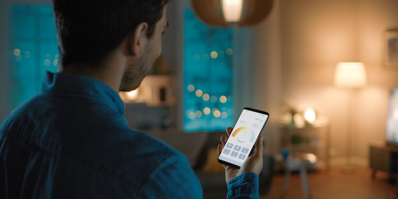 Young Handsome Man is Setting up Comfortable Bedroom Conditions with Voice Command to Smart Home Application on His Smartphone. It's a Cozy Evening in Technologically Advanced Living Room.